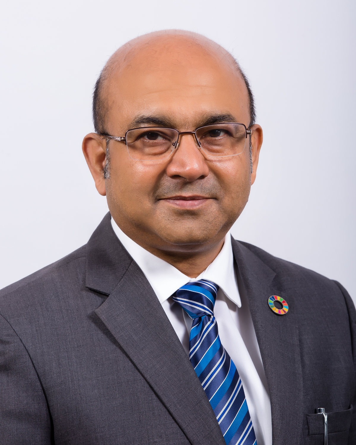 A photo of a man wearing a grey suit, blue tie and glasses.