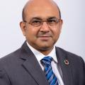 A photo of a man wearing a grey suit, blue tie and glasses.